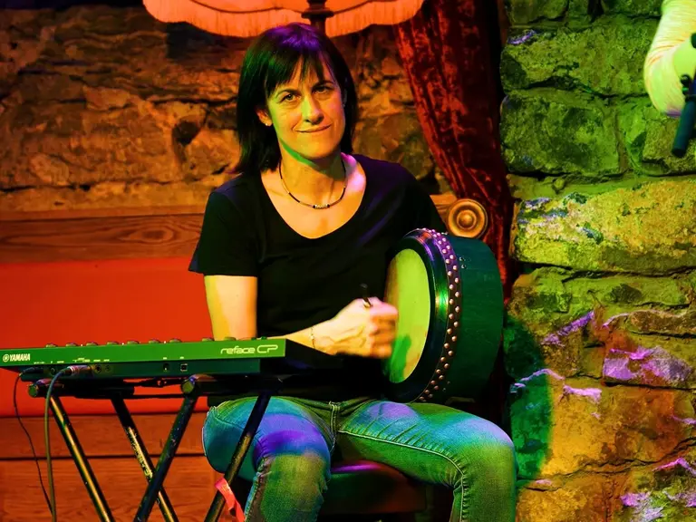 Musikerin spielt eine Bodhrán in einem Pub. Sie wird mit grünen und roten Lichtern angeleuchtet. 