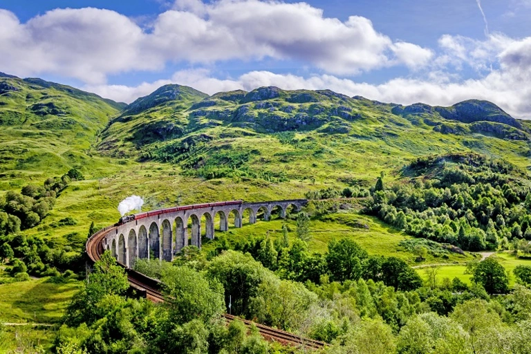 Das Glenfinnan-Viadukt in den schottischen Highlands mit einem vorbeifahrenden historischen Dampfzug, bekannt als „Hogwarts-Express“. Die lange Steinbrücke windet sich durch eine weitläufige, grüne Hügellandschaft unter blauem Himmel mit weißen Wolken.