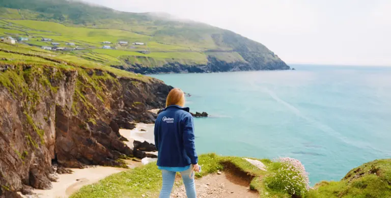 Wandern auf der Dingle-Halbinsel: Irlands spektakuläre Steilküste Eine Frau steht auf einem Küstenpfad der Dingle-Halbinsel in Irland und blickt auf die türkisfarbene See. Unter ihr fallen grüne Klippen steil ab, im Hintergrund liegen verstreute Häuser auf grünen Hügeln unter leichtem Küstennebel.