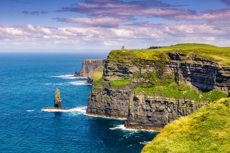 Die Cliffs of Moher an der Westküste Irlands mit steilen, grünen Klippen, die hoch über dem Atlantik aufragen. Das tiefblaue Meer trifft auf markante Felsformationen, während der Himmel in sanften Abendfarben leuchtet.