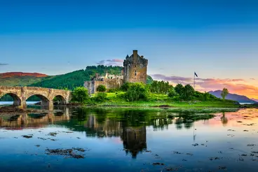 Eilean Donan Castle in den schottischen Highlands, malerisch gelegen auf einer kleinen Insel im Loch Duich. Die historische Burg aus Stein ist über eine Bogenbrücke mit dem Festland verbunden und spiegelt sich im ruhigen Wasser bei Sonnenuntergang.
