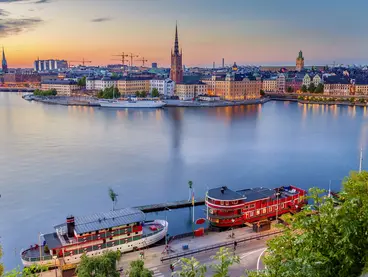 Eine Luftaufnahme der Stadt Stockholm bei Sonnenuntergang mit Blick auf historische Gebäude, Kirchen und Boote im Hafen. Das Wasser spiegelt den farbenfrohen Himmel wider.