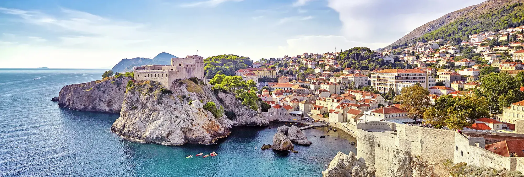 Panorama der historischen Altstadt von Dubrovnik in Kroatien mit mächtigen mittelalterlichen Stadtmauern, der Festung Lovrijenac auf einem Felsen am Adriatischen Meer, roten Ziegeldächern, engen Gassen und den umliegenden grünen Hügeln bei strahlendem Sonnenschein.