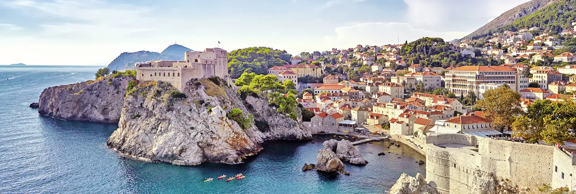Panorama der historischen Altstadt von Dubrovnik in Kroatien mit mächtigen mittelalterlichen Stadtmauern, der Festung Lovrijenac auf einem Felsen am Adriatischen Meer, roten Ziegeldächern, engen Gassen und den umliegenden grünen Hügeln bei strahlendem Sonnenschein.
