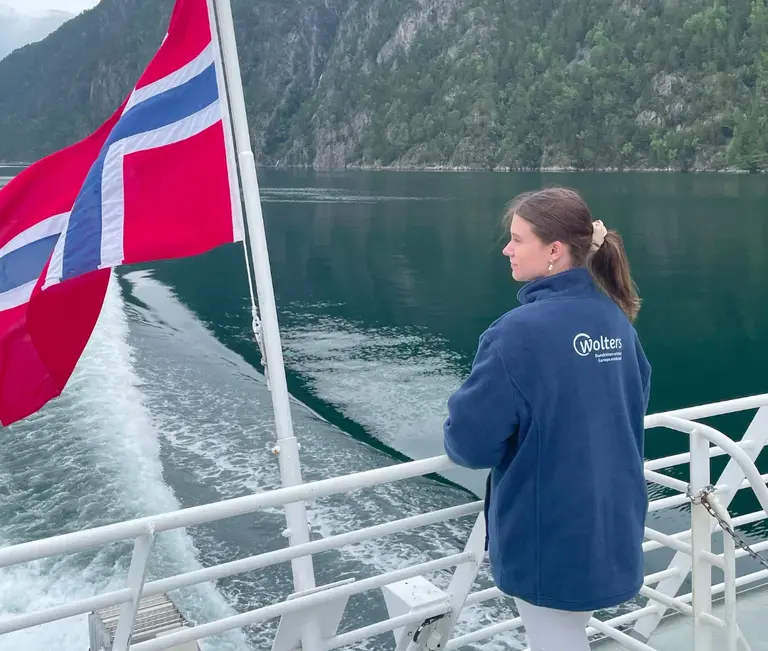 Fjordfahrt Geiranger Auf diesem Bild ist eine Frau zu sehen, wie sie an Deck einer Fähre steht und in die beeindruckende Landschaft eines norwegischen Fjords blickt. Neben ihr weht die norwegische Flagge im Wind, während das Schiff sein Kielwasser durch das ruhige, von steilen, bewaldeten Hängen umgebene Wasser zieht.