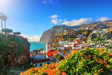 Madeiras Küstenidylle: Farbenfrohes Dorf mit Blick auf den Atlantik Blick auf ein farbenfrohes Küstendorf auf Madeira, eingebettet zwischen steilen Felsklippen und dem türkisblauen Atlantik. Im Vordergrund leuchten orangefarbene Blüten, dahinter steigen Häuser terrassenförmig den Hang hinauf unter einem klaren, sonnigen Himmel.