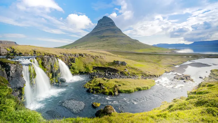 Der markante pyramidenförmige Berg Kirkjufell erhebt sich hinter den Wasserfällen Kirkjufellsfoss; klare Bäche, moosgrüne Wiesen und weitläufige Fjordlandschaft unter blau-weißen Wolken – ikonisches Fotomotiv der Halbinsel Snæfellsnes.