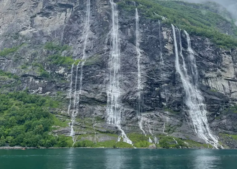 Wasserfall „Die Sieben Schwestern“ Auf diesem Bild ist der berühmte Wasserfall „Die Sieben Schwestern“ (De syv søstrene) zu sehen, wie er im Geirangerfjord in Norwegen eine steile Felswand hinabstürzt. Man erkennt die sieben einzelnen Ströme, die aus großer Höhe direkt in das türkisfarbene Wasser des Fjords fallen und ein spektakuläres Naturdenkmal bilden.