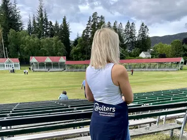 Ein Tag bei den Highland Games: Schottlands Kultur hautnah erleben Eine Besucherin blickt von den Zuschauerrängen auf das grasbewachsene Wettkampffeld eines Highland-Games-Parks in Schottland, umgeben von der malerischen Natur der Highlands, und taucht ein in die faszinierende Kultur und Tradition des Landes während ihres Urlaubs.