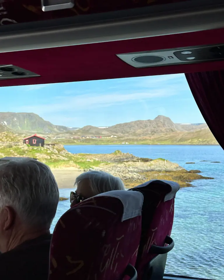 Busfahrt durch die Arktis Auf diesem Bild ist die Aussicht aus einem Reisebus auf die karge und felsige Küstenlandschaft Nordnorwegens zu sehen. Man erkennt ein einzelnes, dunkles Haus an der Küste eines arktischen Fjords, umgeben von baumlosen Bergen, was die Weite und Abgeschiedenheit der Region unterstreicht.