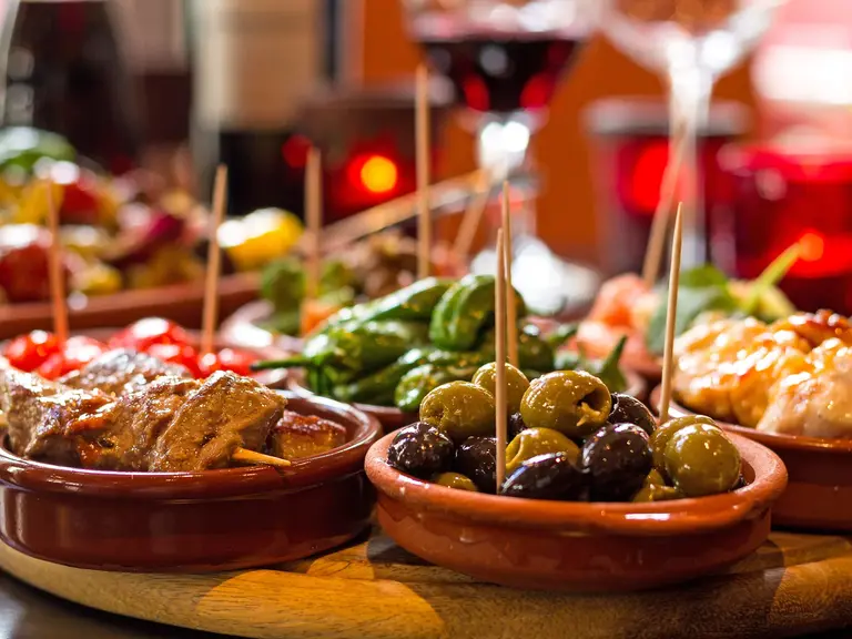 Eine Auswahl klassischer Tapas wie Oliven, Fleischspieße und Paprika steht auf einem Holztisch, begleitet von Rotwein und warmen Licht. Das Bild fängt die gesellige Atmosphäre spanischer Bars ein und steht für die kulinarische Vielfalt und Lebensfreude, die Spanien zu einem beliebten Reiseziel für Genießer macht.