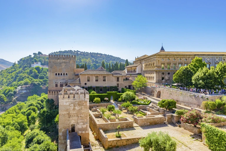 Alhambra, Granada Blick auf die Alhambra in Granada mit Türmen, Mauern und umliegenden Hügeln unter klarem Himmel.