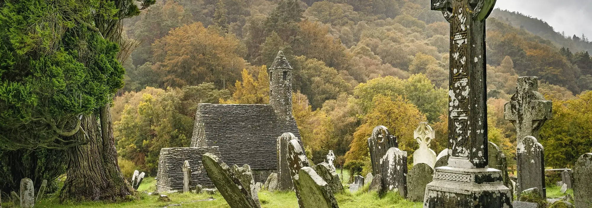 Historische Klosterruine Glendalough in Irland mit keltischen Hochkreuzen, alten Grabsteinen und herbstlich gefärbtem Wald – beliebte Sehenswürdigkeit in den Wicklow Mountains.