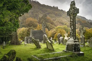 Historischer Friedhof in Glendalough, Irland, mit einem kunstvoll verzierten keltischen Hochkreuz aus Stein im Vordergrund. Umgeben von alten Grabsteinen, Ruinen einer Klostersiedlung und herbstlich gefärbten Bäumen im Wicklow Mountains Nationalpark.