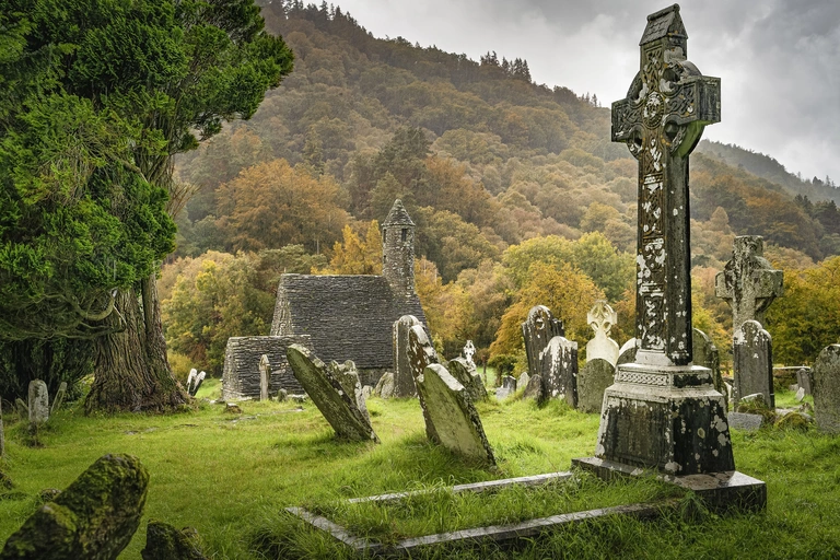 Historischer Friedhof der Klostersiedlung Glendalough in Irland mit alten keltischen Grabkreuzen und verwitterten Grabsteinen. Die mittelalterlichen Ruinen liegen in einem grünen Tal, umgeben von herbstlich gefärbten Bäumen und nebliger, stimmungsvoller Landschaft.