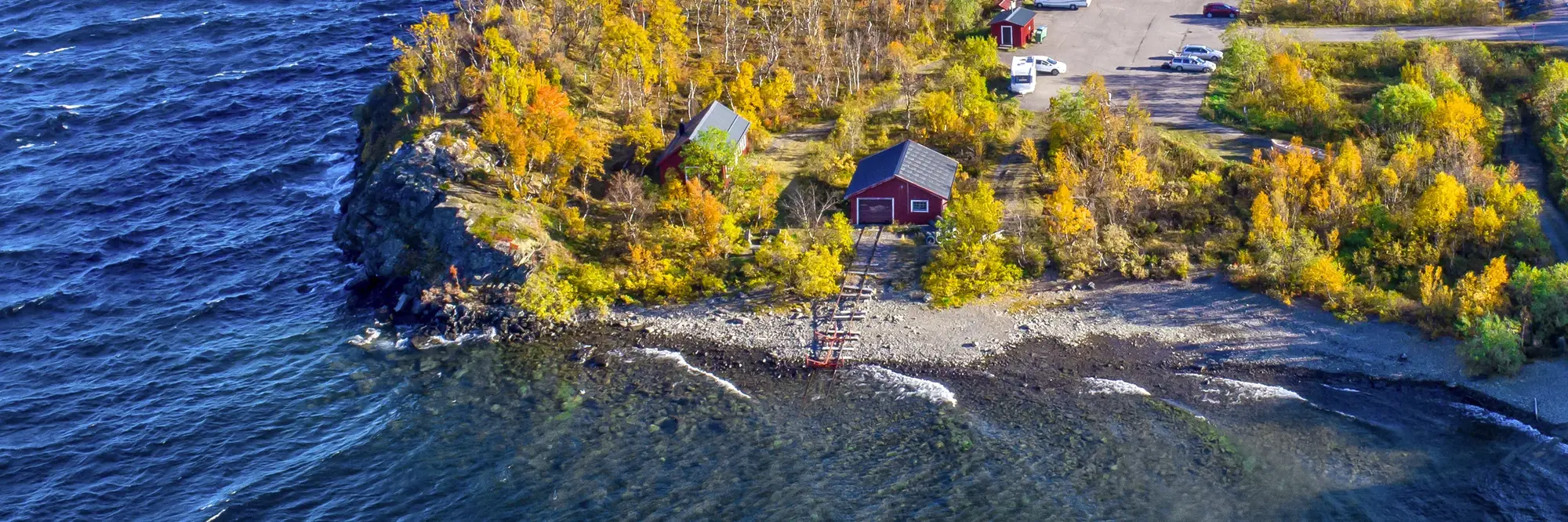 Straße entlang des tiefblauen Torneträsk-Sees in Nordschweden mit Blick auf schneebedeckte Berge. Bunte Herbstbäume und klares Licht schaffen eine beeindruckende Lappland-Landschaft.