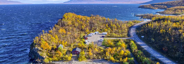Straße schlängelt sich entlang des tiefblauen Torneträsk-Sees in Nordschweden. Herbstfarben leuchten, im Hintergrund liegen schneebedeckte Berge.