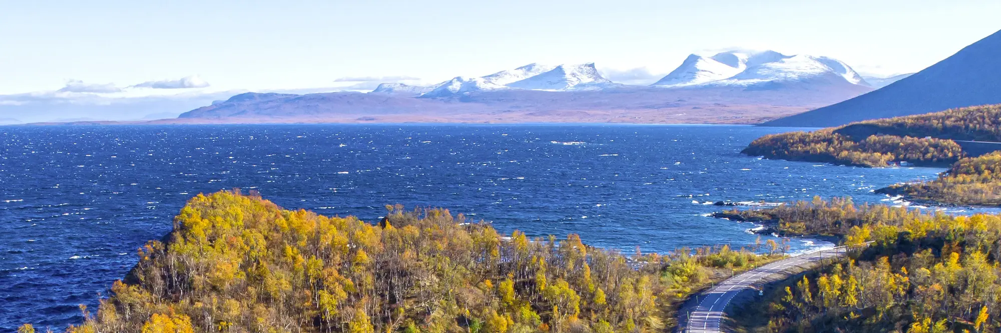 Straße entlang des tiefblauen Torneträsk-Sees in Nordschweden mit Blick auf schneebedeckte Berge. Bunte Herbstbäume und klares Licht schaffen eine beeindruckende Lappland-Landschaft.
