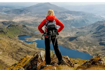 Eine Wanderin mit Rucksack steht auf einem Gipfel und blickt über eine weite Berglandschaft mit Seen und Tälern. Die felsige Anhöhe im Vordergrund führt den Blick in die hügelige Natur unter klarem Himmel – ideal für Outdoor- und Wanderurlaub.