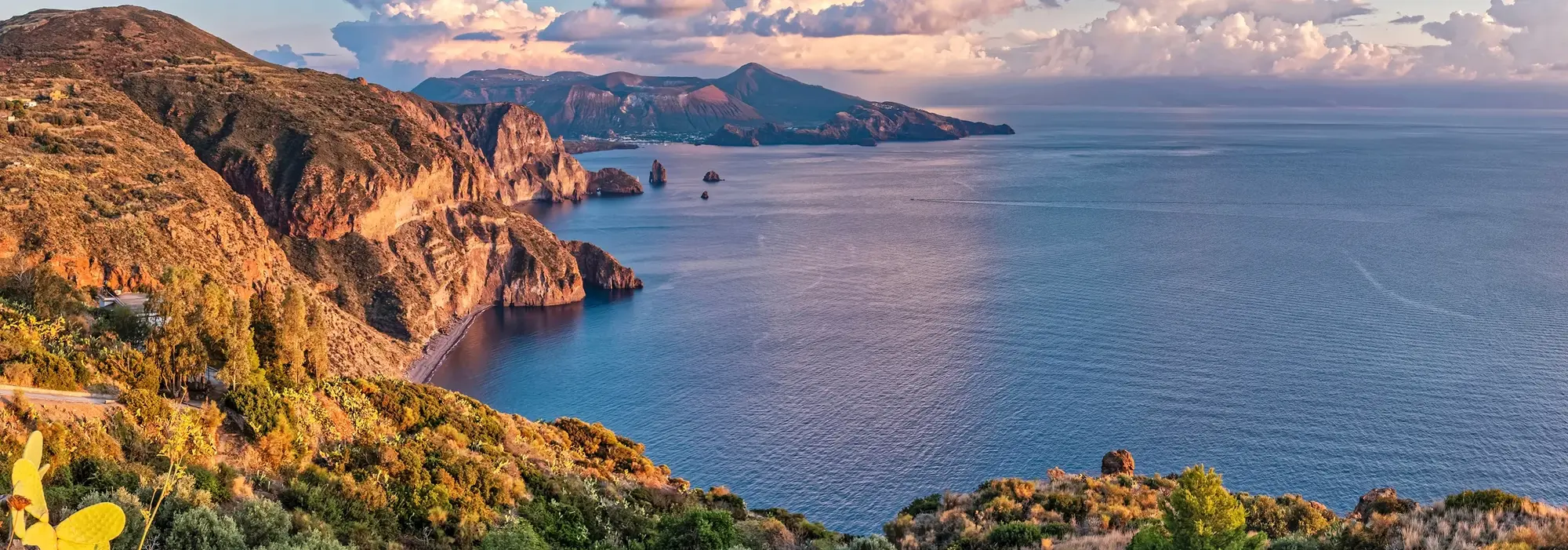 Blick auf die Küste von Lipari, eine der Äolischen Inseln in Italien, mit roten Felsen, tiefblauem Meer und weiter Aussicht bis zu den Nachbarinseln.