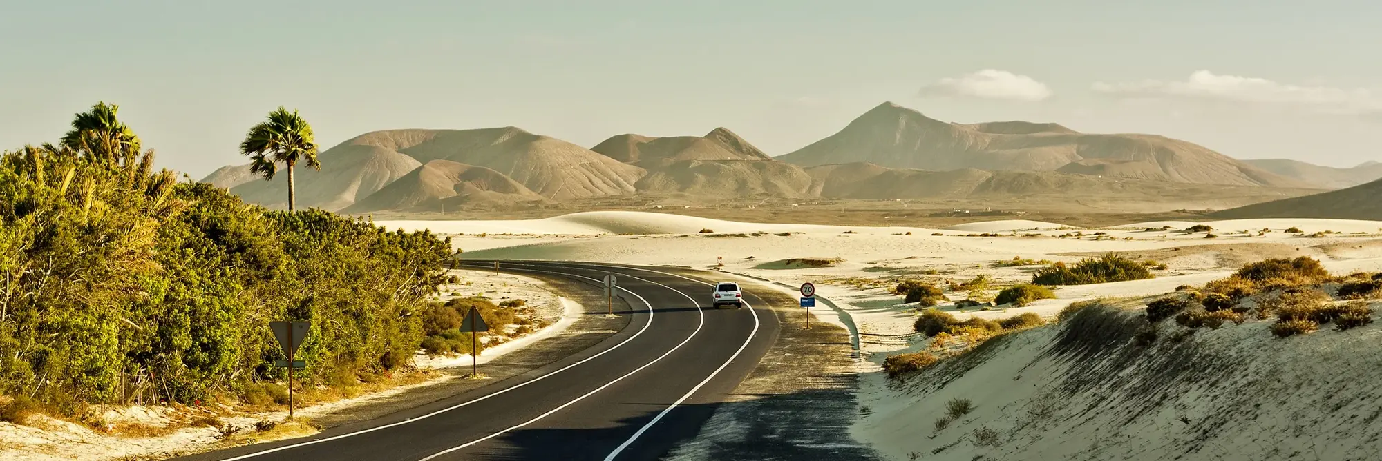 Kurvenreiche Straße durch die Sandlandschaft von Fuerteventura – Natur und Freiheit auf den Kanaren Eine geschwungene Asphaltstraße führt durch eine helle Sand- und Dünenlandschaft auf Fuerteventura auf den Kanarischen Inseln in Spanien, während im Hintergrund sanfte Vulkanberge unter warmem Sonnenlicht aufragen und Palmen sowie karge Vegetation die ruhige, weite Atmosphäre prägen, die typisch für einen Urlaub voller Natur, Reisen, Freiheit und Abenteuer auf der Insel ist.