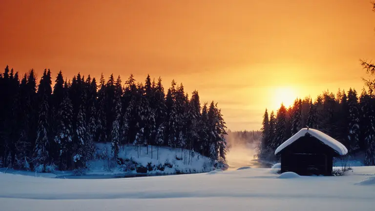 Verschneite Waldlandschaft bei Sonnenuntergang mit Holzhütte und zugefrorenem See – typisch Winteridylle.