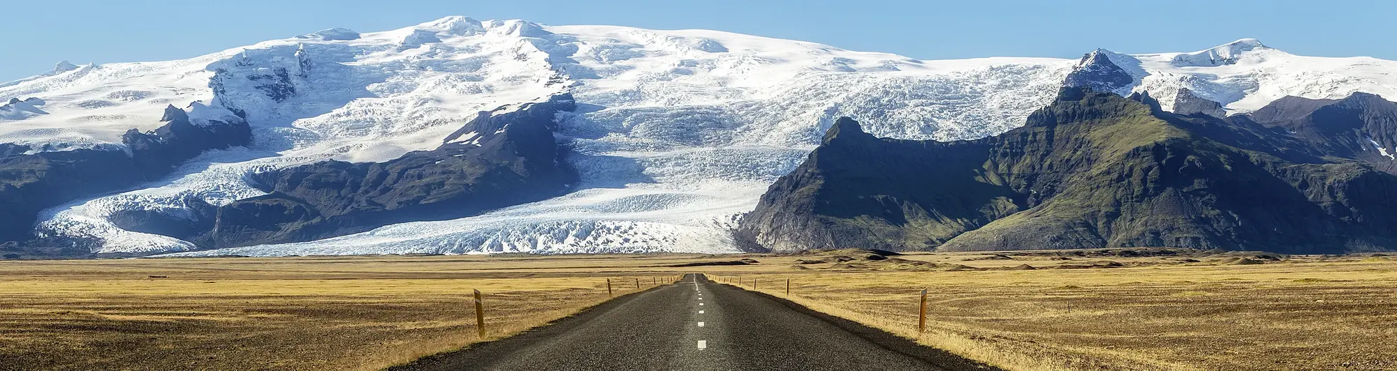 Island-Roadtrip: Endlose Straße vor spektakulärem Gletscherpanorama Eine gerade Landstraße führt durch eine weite, goldbraune Ebene direkt auf einen schneebedeckten Gletscher zu. Im Hintergrund erheben sich dunkle Felsformationen und strahlend weiße Eisflächen unter blauem Himmel.