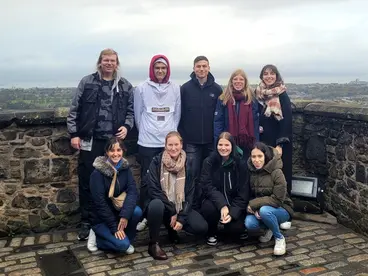 Eine Gruppe von Reisenden steht lachend auf einer alten Steinmauer mit Blick über die Stadt Edinburgh in Schottland. Umgeben von herbstlicher Landschaft vermittelt das Foto die Freude am gemeinsamen Reisen und Erkunden kultureller Highlights während einer Gruppenreise nach Großbritannien.