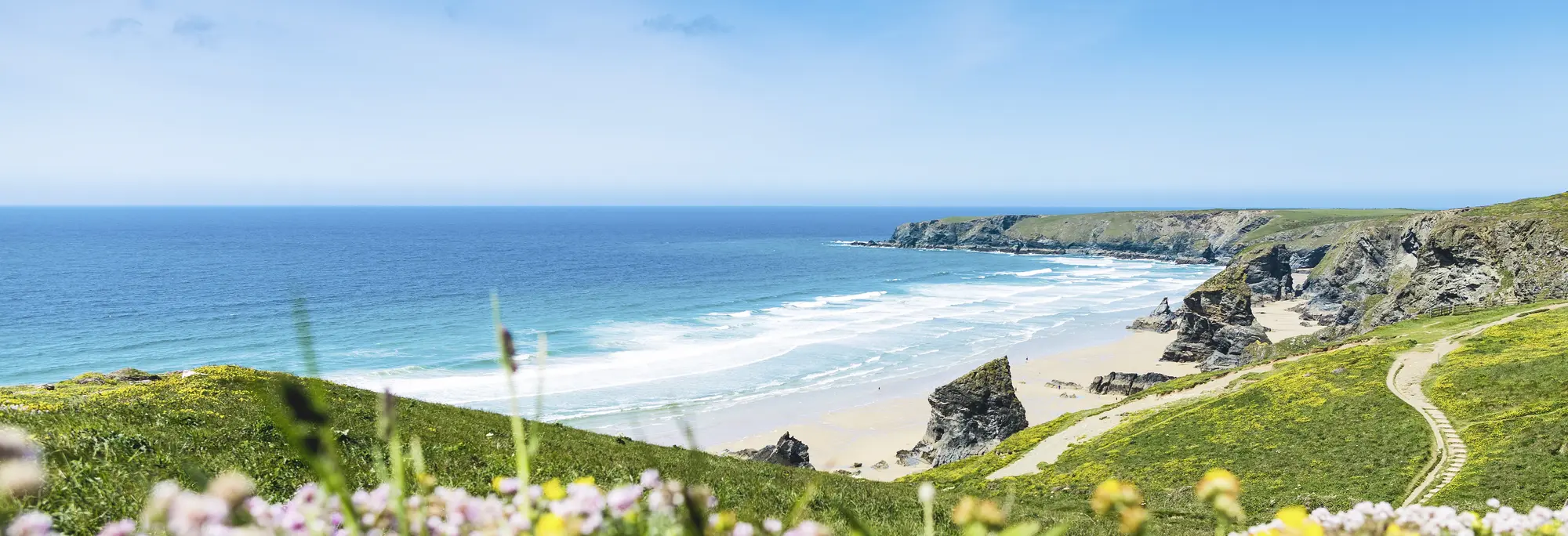 Atemberaubende Küstenlandschaft in Cornwall mit hohen Klippen, türkisfarbenem Meer, feinem Sandstrand und bunten Wildblumen im Vordergrund.