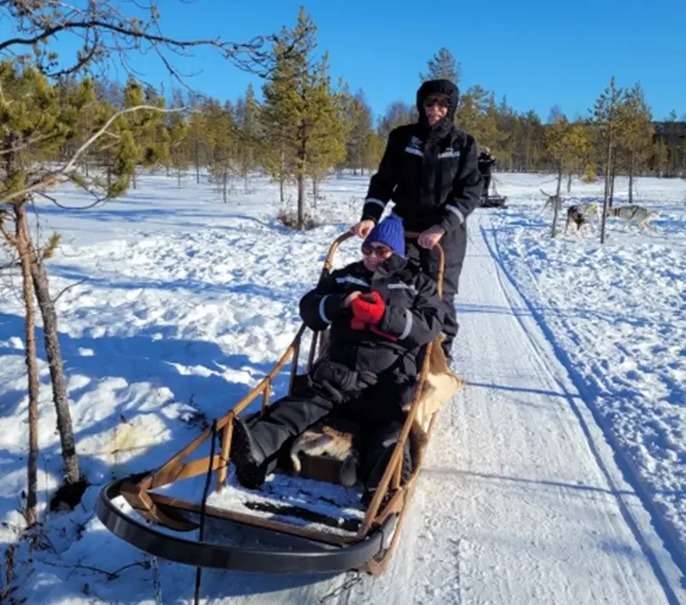 Hundeschlittenfahrt im Schnee In diesem Bild sehen Sie zwei Personen, die an einem sonnigen Wintertag an einer Hundeschlittenfahrt teilnehmen. Eine Person lenkt den traditionellen Holzschlitten, während die andere warm eingepackt darin sitzt und die Fahrt durch die verschneite Landschaft Lapplands genießt.