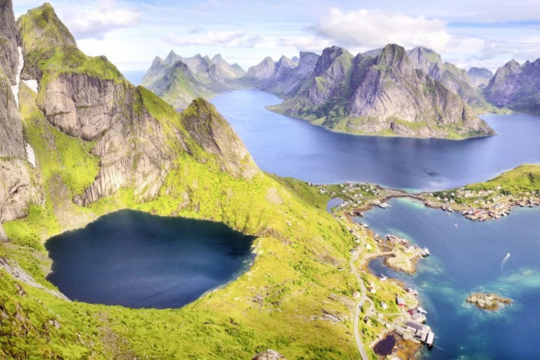 Lofoten in Norwegen – Spektakuläre Inselwelt zwischen Bergen und Fjorden Luftaufnahme der Lofoten in Norwegen mit markanten, steil aufragenden Bergen, tiefblauen Fjorden und kleinen Inseln. Ein runder Bergsee liegt eingebettet in grünes Gelände, während sich Küstenorte und klare Gewässer in der sonnigen Landschaft zeigen.