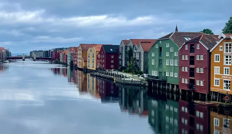 Speicherhäuser am Nidelva Auf diesem Bild ist die ikonische Reihe bunter Speicherhäuser zu sehen, die auf Pfählen am Ufer des Flusses Nidelva in Trondheim stehen. An einem bewölkten, windstillen Tag spiegeln sich die farbenfrohen Fassaden der historischen Gebäude malerisch im ruhigen Wasser des Flusses.
