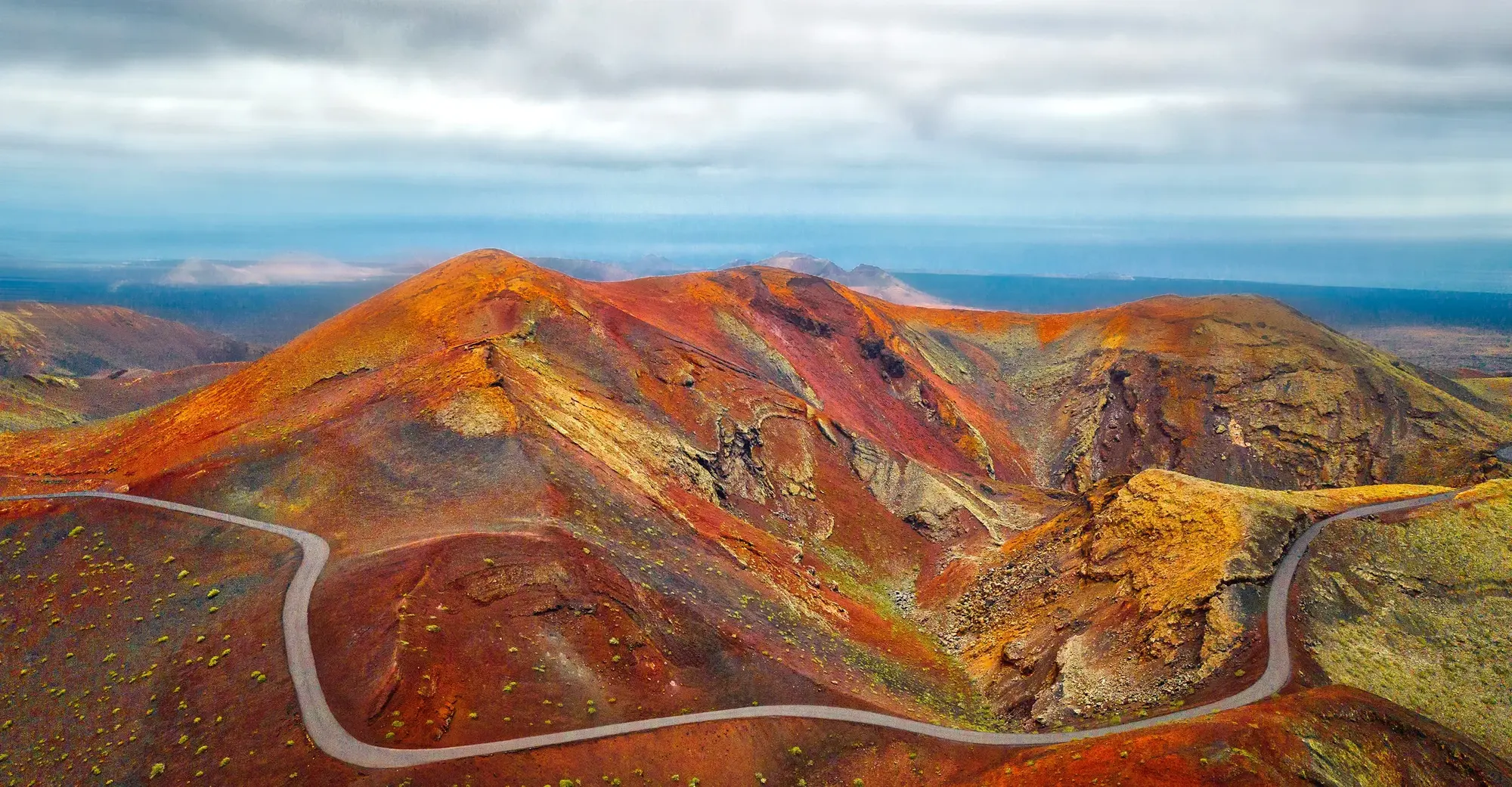 Vulkanlandschaft mit Straße Bergige Vulkanlandschaft mit rötlichem und orangefarbenen Hügeln. Eine kurvige Straße führt durch das Gebiet. Der Himmel ist bewölkt und hinter den Hügeln kann man den Horizont erkennen.