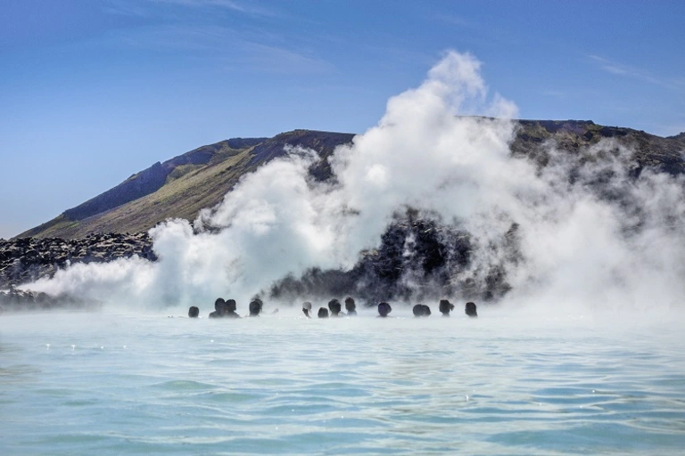Dampfende Blaue Lagune vor vulkanischer Kulisse Blick auf die berühmte Blaue Lagune in Island, in der Badegäste im milchig-blauen Thermalwasser entspannen, während dichte Dampfwolken vor einer vulkanischen Berglandschaft aufsteigen.