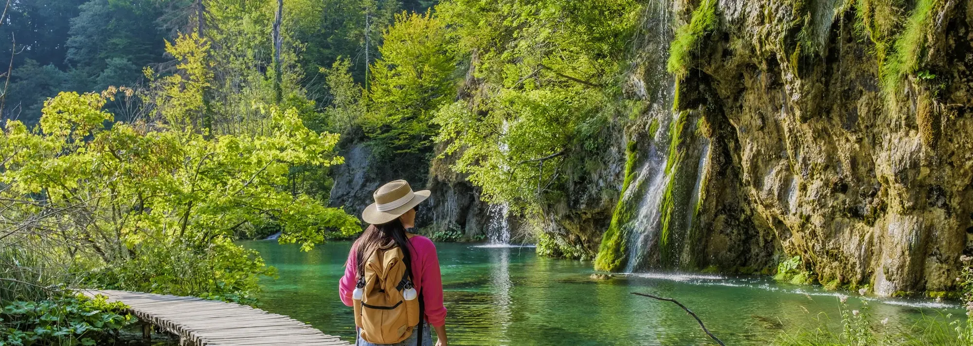 Auf dem Bild sieht man eine Frau mit Hut auf einem Steg im Nationalpark Plitvicer Seen in Kroatien. Das Bild zeigt klares Wasser und einen Wasserfall im Plitvicer Seen Nationalpark im Hintergrund. Ein echtes Highlight auf einer Kroatienreise.