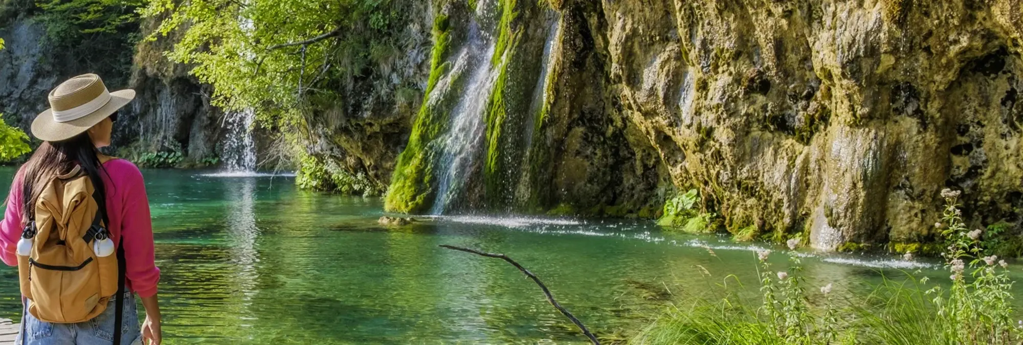Auf dem Bild sieht man eine Frau mit Hut auf einem Steg im Nationalpark Plitvicer Seen in Kroatien. Das Bild zeigt klares Wasser und einen Wasserfall im Plitvicer Seen Nationalpark im Hintergrund. Ein echtes Highlight auf einer Kroatienreise.