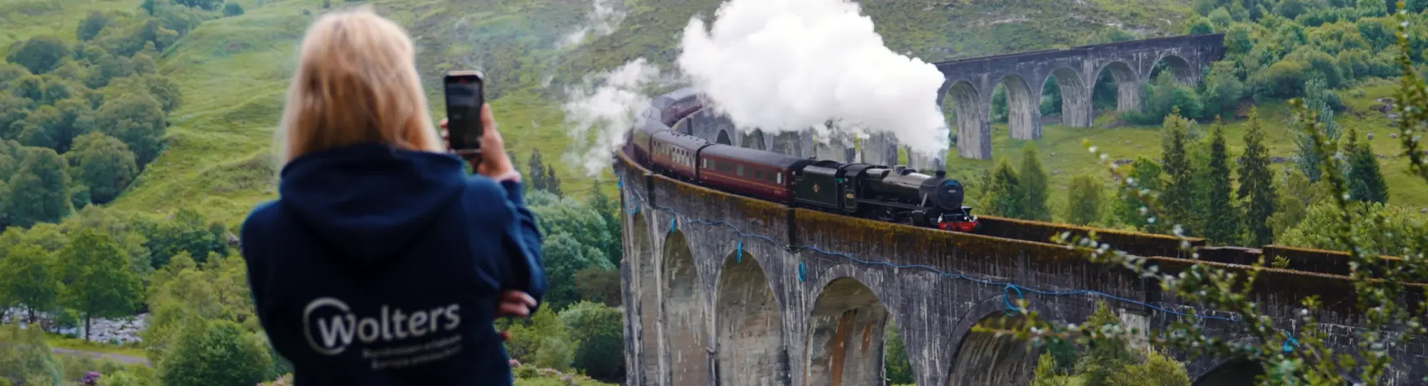 Eine Person fotografiert den berühmten Dampfzug, der über das Glenfinnan Viadukt in den schottischen Highlands fährt. Umgeben von grünen Hügeln und dichter Natur vermittelt die Szene den Zauber klassischer Zugreisen und unvergesslicher Schottland-Erlebnisse.