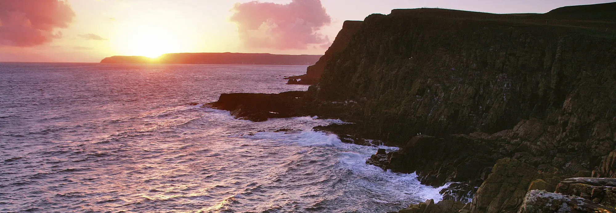 Nordirland Küste - Sonnenuntergang über dem Atlantik Das Bild zeigt die wilde Küstenlandschaft Nordirlands mit steilen Klippen, gegen die die Wellen des Atlantiks schlagen. Die untergehende Sonne taucht den Himmel und das Meer in warme Farben. Typisch für die Region sind die dramatischen Klippenlandschaften und Naturkulissen, die Nordirland zu einem beliebten Ziel für Naturreisen und Roadtrips machen.