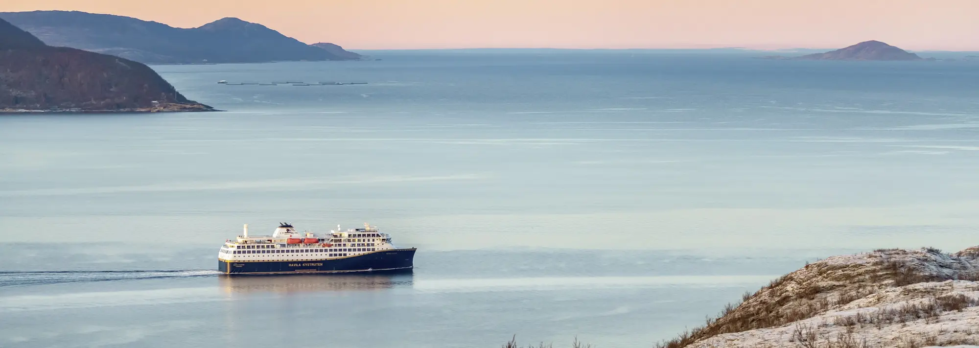 Mit dem Postschiff entlang der Küste: Eine Fjord-Reise in Norwegen Ein modernes Postschiff der Havila Kystruten gleitet bei Sonnenuntergang durch die ruhigen Gewässer der norwegischen Fjordlandschaft und bietet Passagieren auf dieser unvergesslichen Reise einen malerischen Anblick auf die Küste.