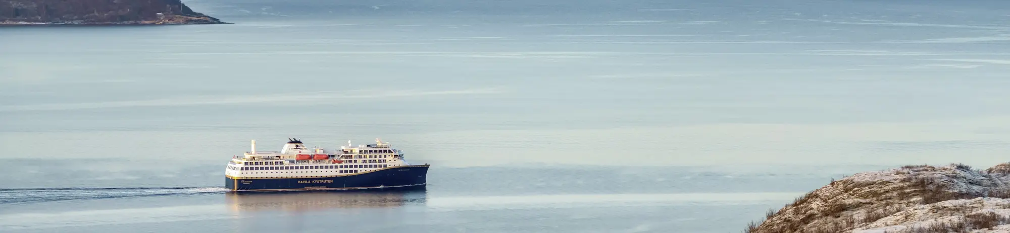Ein modernes Postschiff der Havila Kystruten gleitet bei Sonnenuntergang durch die ruhigen Gewässer der norwegischen Fjordlandschaft und bietet Passagieren auf dieser unvergesslichen Reise einen malerischen Anblick auf die Küste.