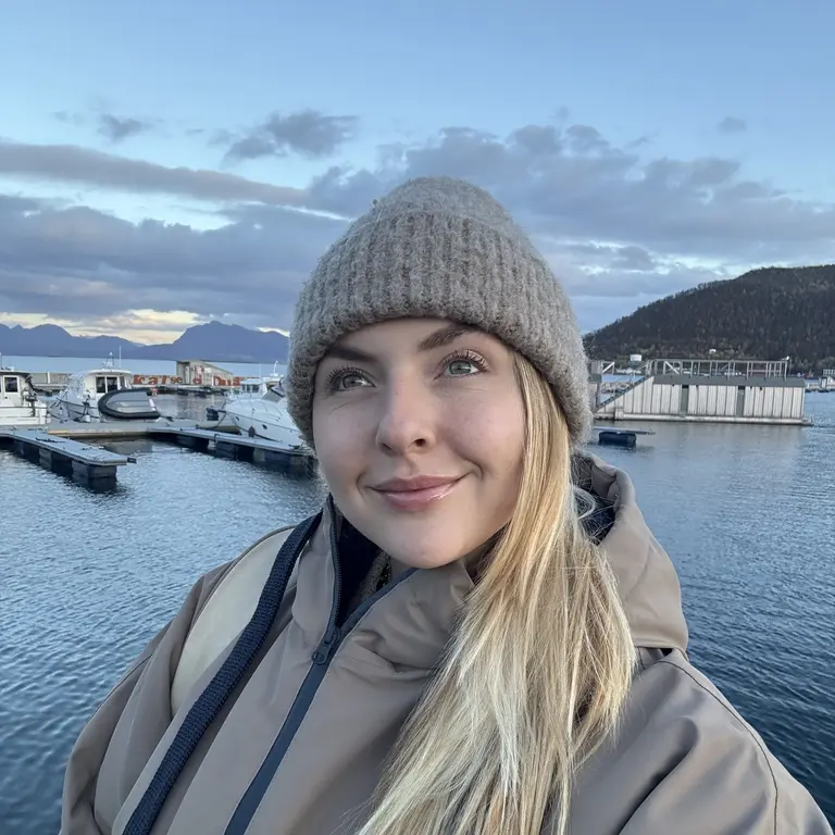 Auf dem Foto steht eine Person in warmer Outdoorbekleidung an einem skandinavisch wirkenden Hafen. Sie trägt eine dicke Strickmütze und eine wetterfeste Jacke, ideal für das kühle Klima Nordnorwegens oder einer ähnlichen Fjordregion. Hinter ihr liegen ruhiges Wasser, kleine Boote, Holzanleger und majestätische Hügel, die in das weiche Licht des frühen Abends getaucht sind. Die Szenerie vermittelt typische Nordland-Atmosphäre: klare Luft, weite Natur, Fjorde und ein stimmungsvoller Himmel – perfekt für Reisethemen rund um Norwegen, Fjordlandschaften oder Outdoor-Abenteuer.