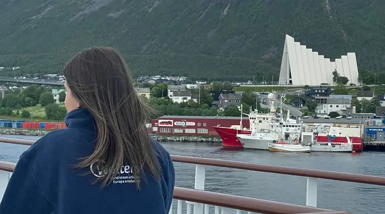 Ankunft in Tromsø Auf diesem Bild ist eine Frau zu sehen, die vom Deck eines Schiffes auf den Hafen von Tromsø blickt. Im Fokus der Ansicht steht die berühmte Eismeerkathedrale (Ishavskatedralen), deren markante, an einen Eisberg erinnernde Architektur sich deutlich von der dahinter liegenden Berglandschaft abhebt.