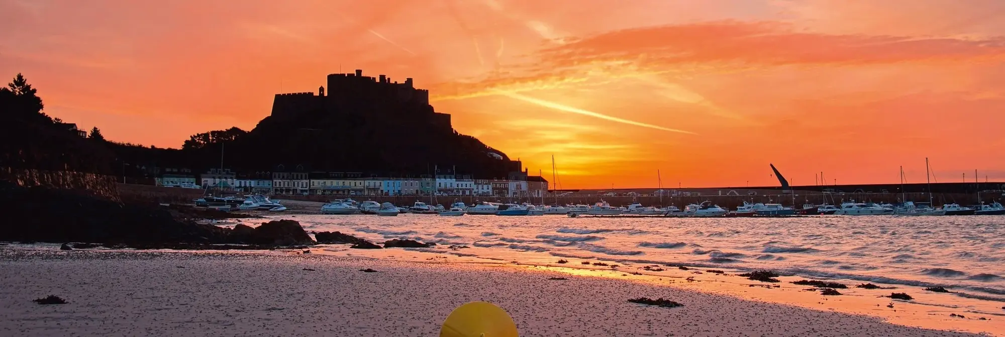Das Mont Orgueil Castle thront als Silhouette über dem Hafen von Gorey auf Jersey. Vor dem Schloss leuchten Fischerboote und Häuser in warmem Abendlicht, während der Himmel in kräftigen Rot- und Orangetönen erstrahlt.
