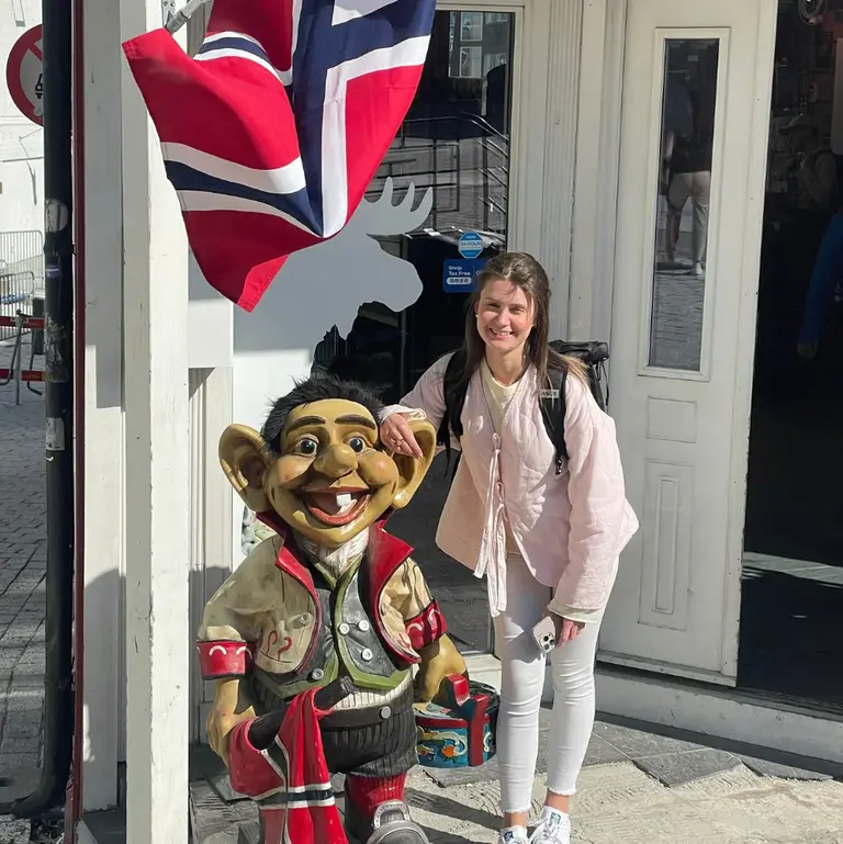 Foto mit einem norwegischen Troll Auf diesem Bild ist eine junge Frau zu sehen, die an einem sonnigen Tag neben einer großen, bunten Troll-Figur vor einem Souvenirladen in Norwegen posiert. Eine norwegische Flagge und eine Elch-Silhouette im Hintergrund unterstreichen das typisch norwegische Urlaubs-Flair der Szene.