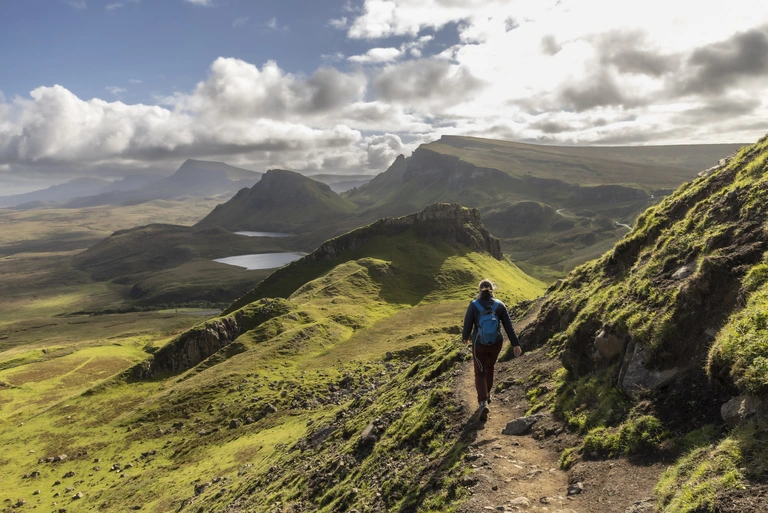 Eine Person wandert auf einem schmalen Pfad durch die schottischen Highlands. Die hügelige, leuchtend grüne Landschaft erstreckt sich bis zum Horizont, mit Seen, Felsformationen und wolkenverhangenem Himmel. Die Szene vermittelt Ruhe, Weite und Naturerlebnis in Schottland.