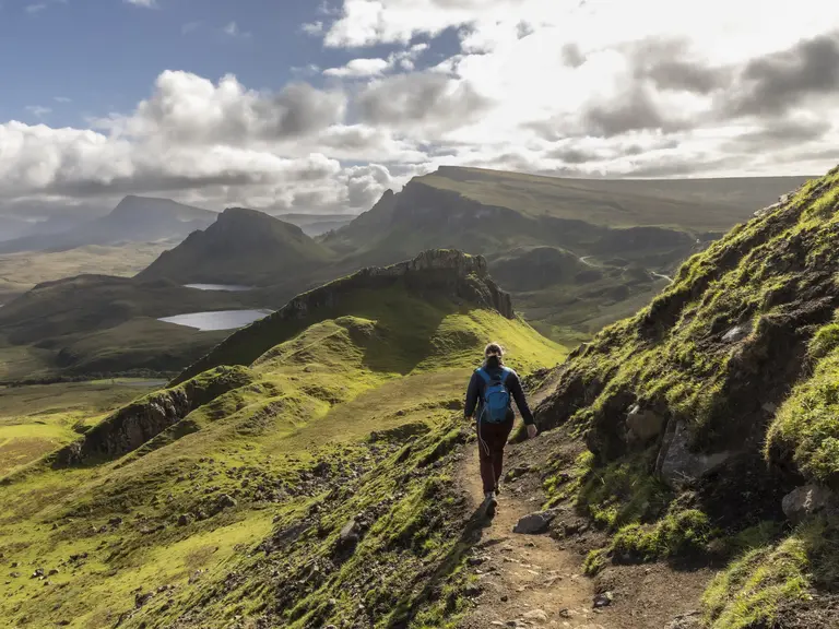 Schottische Highlands – Wanderung durch die wilde Natur Schottlands Eine Person wandert auf einem schmalen Pfad durch die grünen Hügel der schottischen Highlands. Sanfte Berge, weite Täler und dramatische Wolken am Himmel prägen die beeindruckende Landschaft in Schottland.