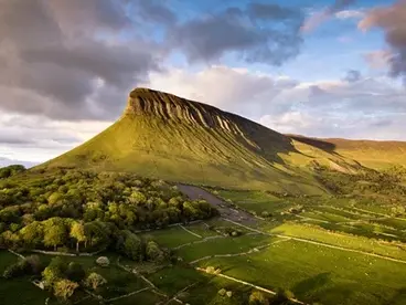 Benbulben in Irland: Wanderung zum majestätischen Tafelberg in Sligo Der majestätische, von der Sonne beschienene Tafelberg Benbulben in der Grafschaft Sligo in Irland ragt bei dramatischem Abendlicht über die saftig grüne Landschaft aus Wäldern und Feldern und lädt als beeindruckende Sehenswürdigkeit zu einer unvergesslichen Reise und einem Wander-Abenteuer in der Natur Irlands ein.