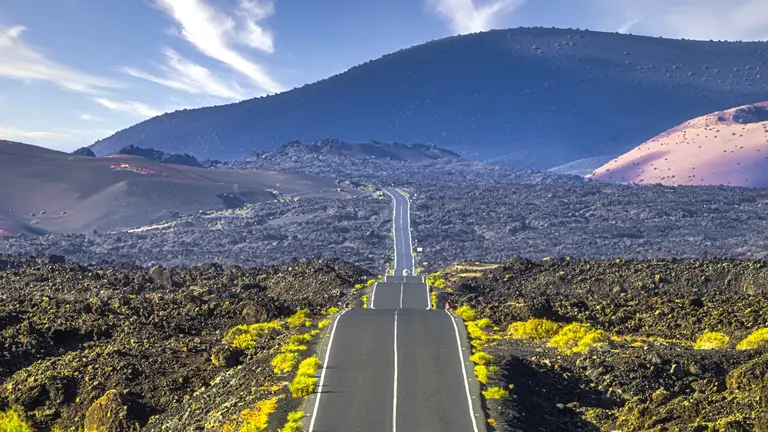 Eine Straße durch eine Vulkanlandschaft Eine Straße verläuft gerade durch eine grüne Vulkanlandschaft. Im Hintergrund erheben sich große Hügel und Berge unter klaren Himmel.