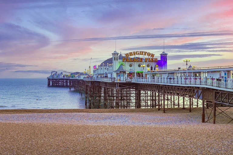 Der Brighton Palace Pier an der Südküste Englands ragt ins Meer hinaus. Die beleuchtete Seebrücke mit Fahrgeschäften und Gebäuden spiegelt sich im Wasser, während die Sonne bei dramatischem Abendhimmel über dem Strand untergeht.