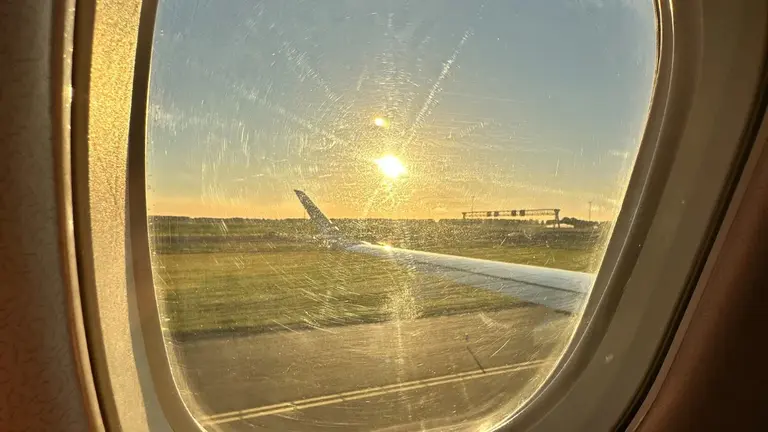 Ankunft in Bremen bei Sonnenaufgang Auf diesem Bild ist der Blick aus einem zerkratzten Flugzeugfenster während des Starts oder der Landung am Flughafen Bremen zu sehen. Man erkennt die Tragfläche des Flugzeugs auf der Rollbahn, während die tiefstehende Morgensonne die Szene in ein helles, goldenes Licht taucht.